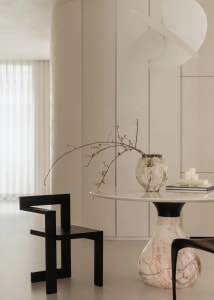 Modern minimalist dining area featuring a round white marble tabletop on a pink marble pedestal base, a black geometric zigzag chair, a textured ceramic vase with dried branches, and a sculptural white pendant lamp against a neutral backdrop.