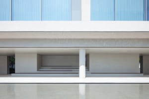 Modern minimalist concrete building facade with large glass windows, white cylindrical column, stepped seating, and a calm reflecting pool in the foreground.