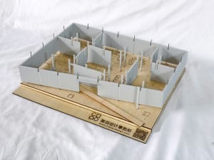Architectural scale model of an interior space with white partition walls, wooden base, and laser-cut furniture layouts, displayed on a white fabric background.