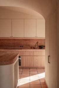 Modern minimalist kitchen featuring terracotta tiles, travertine countertops, light wood cabinetry, and a curved plaster archway with warm natural light.