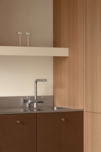 Close-up of a modern kitchen sink area with stainless steel faucet, integrated stainless steel backsplash and sink, warm wood lower cabinets with round pulls, and a floating white shelf above with two glass tumblers.