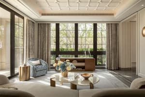 Bright modern luxury living room with large windows overlooking greenery, featuring a blue and white patterned armchair, gold textured side table, sculptural white marble coffee table, and neutral sofa.