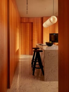 Narrow kitchen hallway with floor-to-ceiling walnut wood cabinetry, white marble kitchen island, two black wooden bar stools, and a large cylindrical pendant lamp.