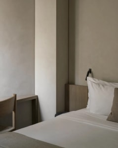 Minimalist bedroom corner featuring a light oak headboard, white bedding, textured beige pillow, black wall sconce, and a partial view of an oak chair against a textured neutral wall.