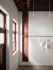 Interior corner of an industrial loft featuring an exposed red brick wall, white painted surfaces, wooden ceiling beams, a metal clothing rail with white garments, and natural light from a wooden window.