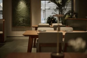 Modern minimalist restaurant dining area featuring light oak wooden tables, beige upholstered chairs, large textured green artwork on the wall, and abundant indoor plants near large windows.