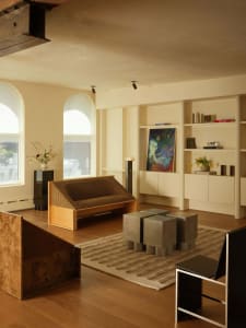 Modern living room featuring a raised burl wood platform, a corduroy-upholstered wooden sofa, four concrete cube stools on a striped rug, built-in shelving, and arched windows.
