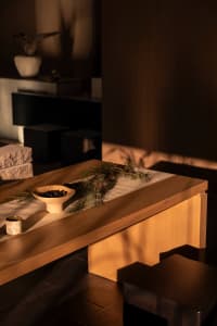 Close-up of a minimalist solid wood dining table featuring a white ceramic bowl with dark berries, pine branches, and a small textured cup under warm dramatic lighting.