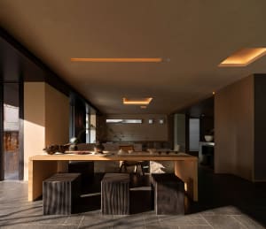 Minimalist dining room featuring a long solid oak table, three dark charred-wood block stools, a woven rattan chair, and large picture windows allowing natural light to flood the space with warm earthy tones.