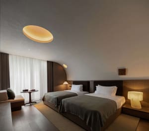 Modern minimalist hotel twin bedroom featuring two beds with dark walnut headboards, warm wood tones, a large circular pendant light, and curved architectural ceiling details.