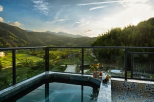 Corner rooftop jacuzzi with dark marble surround and glass railing overlooking lush green mountain valley at sunset