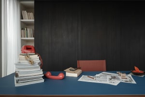 Blue desk with stacks of books, a red vintage rotary telephone, scattered newspapers, a magnifying glass, and a red leather chair in front of dark wood paneling and a built-in bookshelf.