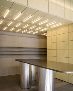 Modern reception desk featuring a curved dark countertop supported by cylindrical stainless steel bases, set against pale yellow perforated wall and ceiling panels with integrated linear lighting.