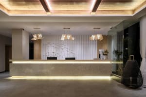 Modern hotel reception desk made of travertine stone with under-lighting, woven rattan pendant lamps, white pleated wall, metallic bird wall art, and wooden ceiling.