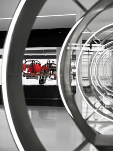 View through large circular steel and glass frames into a modern exhibition hall displaying red car chassis and mechanical components on a white platform.