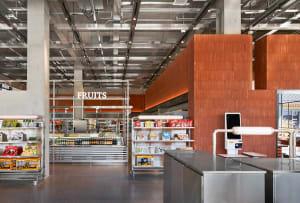 Modern supermarket interior showing stainless steel checkout counters, orange-brown corten steel feature walls, metal shelving units, and an exposed industrial ceiling with visible ductwork and lighting.