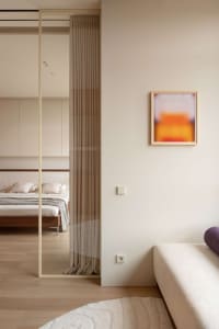 Modern minimalist bedroom viewed through a walnut-framed sliding glass partition with pleated curtain, featuring a low wooden bed with white bedding and abstract orange artwork on the adjacent wall.