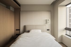 Modern minimalist bedroom with a beige upholstered bed, white linens, vertical wood panel accent wall, and large window with natural light.
