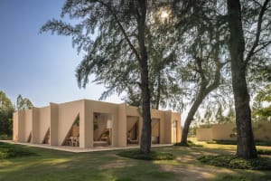 Exterior view of a modern beige villa featuring angular sandstone walls and large openings, surrounded by lush tropical trees and manicured lawn under a clear blue sky.