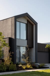 Modern two-story house exterior featuring dark grey stucco, vertical dark wood cladding, large windows with sheer curtains, and landscaped garden with native plants under a clear blue sky.