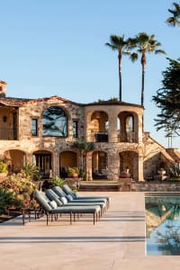 Luxurious Mediterranean-style villa with rough stone walls, arched windows, red tile roof, teal outdoor lounge chairs on a travertine patio, and a swimming pool surrounded by palm trees under a clear blue sky.