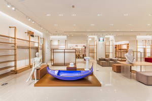 Modern fashion boutique interior featuring a glossy blue Venetian gondola display on a wooden platform, surrounded by minimalist oak shelving and white mannequins under bright track lighting.