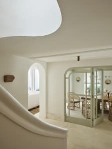 View from a curved staircase into a bright dining room with sage green arched French doors, rattan dining chairs, and white plaster walls.