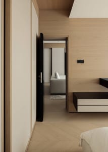 Modern minimalist bedroom interior featuring light oak wood paneling, herringbone wood flooring, built-in dark wood nightstand, and a view into an adjacent seating area.