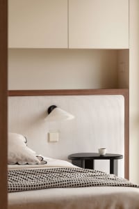 Modern minimalist bedroom with a cream-colored vertically ribbed fabric headboard, black cone-shaped wall sconce, dark oval bedside table, and neutral bedding with black-and-white checkered throw.