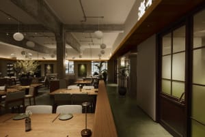 Interior view of a modern Japanese restaurant featuring long light oak dining tables, upholstered chairs, exposed concrete beams, and hanging spherical pendant lights.