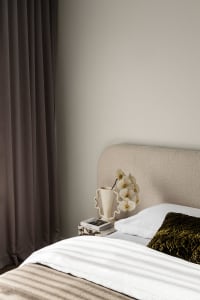 Modern minimalist bedroom corner featuring a textured beige fabric headboard, white bedding, an olive green velvet quilted pillow, and a marble nightstand with a sculptural white vase holding white orchids.