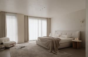 Minimalist bedroom with a large upholstered bed dressed in beige linen, a curved white bouclé sofa, sheer curtains allowing soft daylight, and a wooden side table with candles.