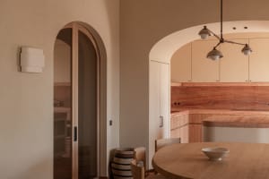 Warm minimalist kitchen and dining area featuring rounded wooden dining table, light oak cabinetry, terracotta marble backsplash, and dramatic arched doorways under a modern multi-arm pendant light.
