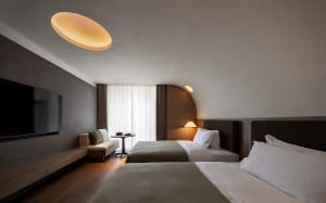 Modern minimalist twin hotel room with two beds, textured grey plaster walls, wooden flooring, built-in TV console, and warm pendant lighting.