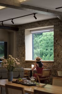 Woman sitting at a wooden dining table reading in front of a large window with a lush green forest view, surrounded by exposed stone walls and wooden furniture.