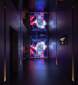 Modern dark hallway with vertical LED lighting strips and two large digital art screens showing colorful abstract fireworks or light explosion patterns in blue, red, and white.