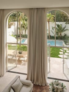 View from a bright sunroom with beige linen curtains, arched glass doors, and outdoor patio furniture overlooking a swimming pool and tropical garden.