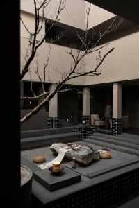 Modern minimalist courtyard with large natural stone slab table, woven cushions, checkerboard tile columns, and a bare olive tree in a sunken stone-paved space.
