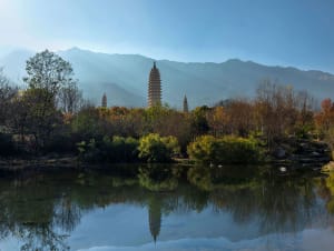 Three ancient pagodas reflected in the calm water of a lake surrounded by autumn trees and distant mountains under a clear blue sky.