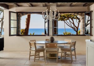 Round dining table with four cane-back chairs positioned in front of large arched windows opening to a bright blue ocean view, white chandelier hanging from beamed ceiling, limestone tile floor.