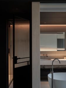 Modern minimalist bathroom featuring raw concrete walls, a white freestanding bathtub with a curved floor-mounted faucet, a stone floating vanity, and a black-framed ribbed glass door with warm backlighting.