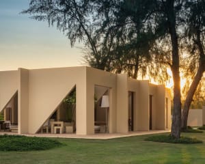 Modern minimalist concrete villa with angular walls and integrated outdoor seating areas surrounded by lush tropical trees at sunset.