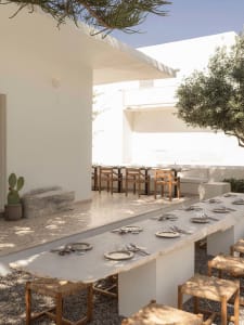 Outdoor dining terrace with a long irregular travertine table, wooden woven stools, set against white minimalist architecture under bright sunlight.