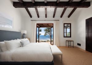 Bright coastal bedroom with large bed dressed in white striped linens, dark wood ceiling beams, and open French doors revealing a stunning ocean view.