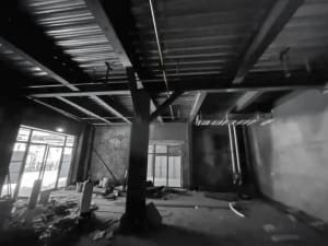 Black and white photograph of an unfinished industrial loft interior showing exposed steel beams, concrete ceiling, structural columns, and large windows with construction debris on the floor.