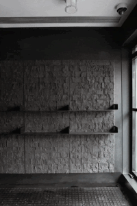 Monochrome interior featuring a textured stacked stone wall with three black floating shelves, dark tiled floor, and industrial-style lighting fixtures.