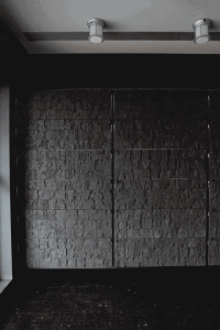 Dark entryway featuring a large textured stacked stone wall with vertical metal dividers and two cylindrical ceiling lights.