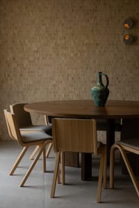 Modern minimalist dining area with a round oak table, four matching oak dining chairs with grey upholstery, a green ceramic pitcher, and a warm brown textured tile wall with two circular wall sconces.