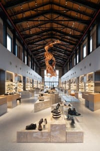 Long view down a modern shoe store with travertine display blocks, wooden floating sculpture, industrial ceiling, and extensive shelving.
