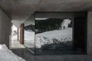 Interior view of a modern concrete mountain house looking out onto a snow-covered alpine forest and ski slope.